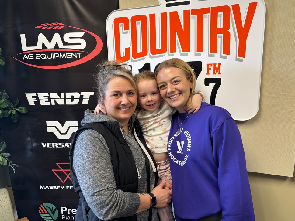 Two women and a young girl standing in front of signs for Country 107 and LMS Ag Equipment.